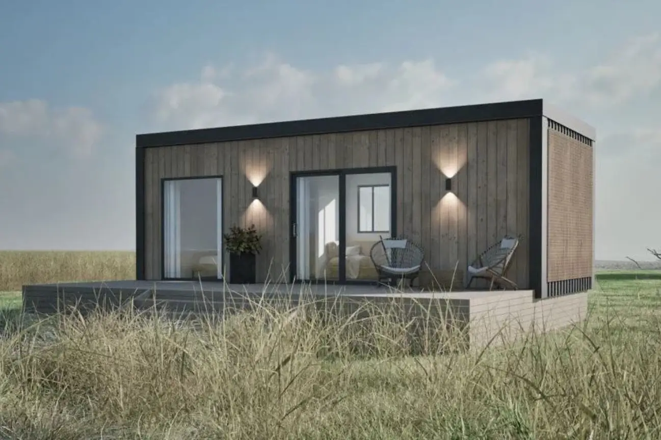 At Tiny Homes New Zealand a sleek timber clad cabin with large sliding doors outdoor deck seating and subtle exterior lighting set in an open grassy landscape