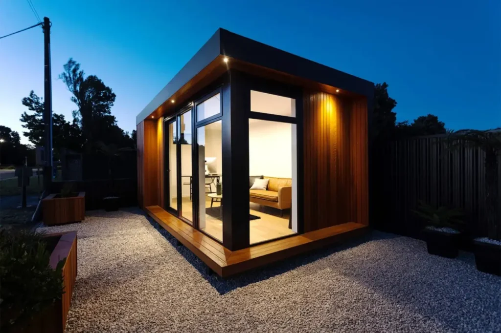 At Tiny Homes New Zealand a warm illuminated studio cabin with timber cladding floor to ceiling windows and a minimalist outdoor setting at dusk