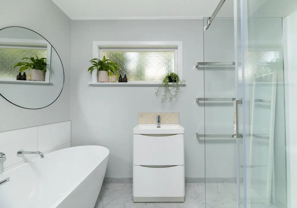 Dark toned modern bathroom with floating wooden vanity round mirror glass shower and textured tiles representing bathroom renovations new zealand