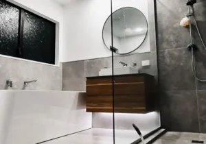Dark toned modern bathroom with floating wooden vanity round mirror glass shower and textured tiles representing bathroom renovations new zealand