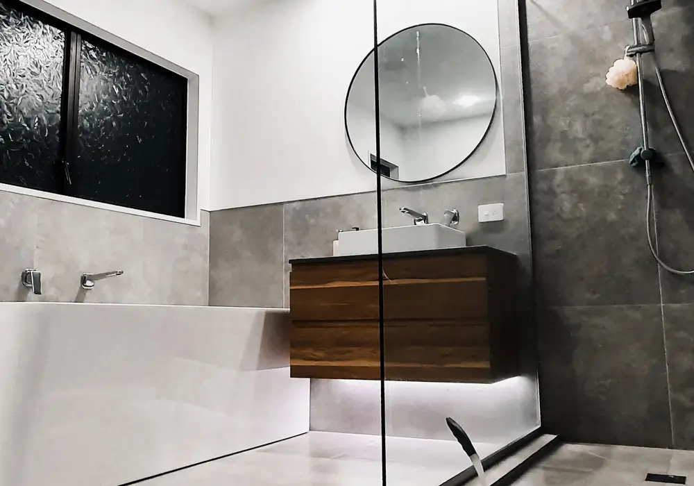 Dark toned modern bathroom with floating wooden vanity round mirror glass shower and textured tiles representing bathroom renovations new zealand