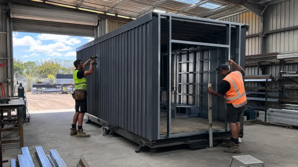 Two Auckland workers in hi vis vests assemble a durable dark grey portable cabin in a well lit workshop using a power drill and level.